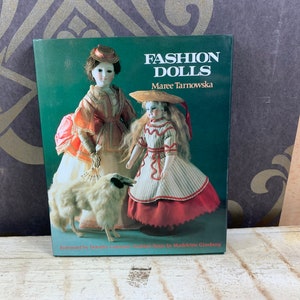 May include: A book titled "Fashion Dolls" by MaRee Tarnowska. The cover features two antique dolls dressed in elaborate gowns and a white dog.