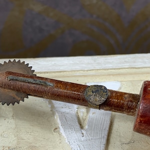 Puede incluir: Un cortador rotatorio vintage de madera y metal con una cuchilla circular y un mango de madera. El mango tiene una banda metálica con el texto "MADE IN ENGLAND" en él.