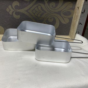 Puede incluir: Tres fiambreras rectangulares de aluminio plateado con asas plegables apiladas sobre una superficie blanca. Las fiambreras tienen un diseño sencillo. El fondo incluye un panel decorativo con un patrón dorado.
