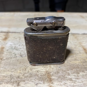 May include: A vintage lighter with a silver metal top and a dark brown leather-wrapped body. The lighter has a rectangular shape and is resting on a wooden surface. The metal top features a textured design for striking.