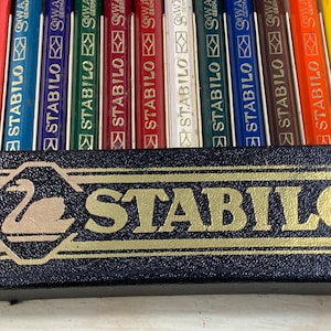 May include: A set of 12 colored pencils in a black box with a gold swan logo and the word "STABILO" printed in gold letters. The pencils are labeled "STABILO" and "GERMAN" in white lettering.