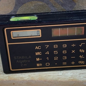 Puede incluir: Una calculadora negra con un nivel de burbuja en la parte superior. La calculadora tiene un borde dorado y una pantalla digital. Los botones están etiquetados con números, símbolos matemáticos y funciones. El texto "STABILA Algebra 2 m" está impreso en la calculadora.
