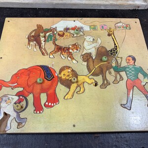 Puede incluir: Un rompecabezas de madera vintage que representa una escena de circo con un maestro de ceremonias, un elefante, un león, un tigre, una jirafa, un camello y un oso polar. Las piezas del rompecabezas están recortadas y tienen pomos para facilitar su manejo.