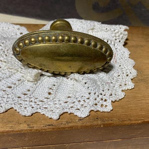 Op de afbeelding: Een antieke ovale deurknop van messing met een kralenrand. De knop ligt op een wit kanten kleedje, op een houten oppervlak. Het verouderde uiterlijk van de knop suggereert vintage charme.