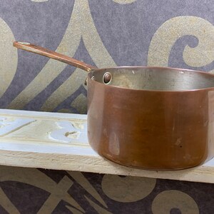 Op de afbeelding: Een kleine koperen pan met een houten handvat. De pan heeft een glanzende, licht aangetast oppervlak.