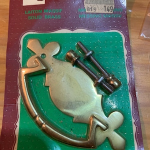May include: A gold-toned, ornate, decorative door handle with a loop design. The handle is packaged on a green and brown card with the text "LAITON MASSIF SOLID BRASS" and "MESSING MASSIV" in French and German. The package also includes two screws.