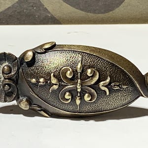 Könnte beinhalten: Antiker Schubladengriff aus Bronze mit detailliertem, symmetrischem Design. Der Griff hat ein zentrales Blumenmotiv und dekorative Schnörkel, mit einer strukturierten, gealterten Oberfläche. Die Gesamtform ist länglich und oval, mit einem abgerundeten Ende und einem fächerförmigen Detail.
