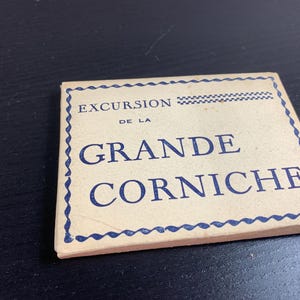 Puede incluir: Una tarjeta rectangular vintage con las palabras "EXCURSION DE LA GRANDE CORNICHE" en letras azules. La tarjeta tiene un borde azul decorativo y está sobre una superficie oscura. La tarjeta parece envejecida.