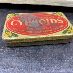 Puede incluir: Una caja de hojalata vintage con una etiqueta roja y dorada que dice "CYPHOIDS - Aceite líquido y extracto o glóbulos - Preparado por Smith & Co. 132, Borough S.E. Londres, Inglaterra."
