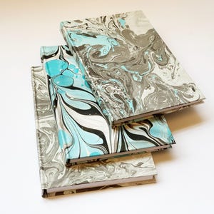 May include: Three notebooks with marbled covers in shades of blue, black, and gray. The notebooks are stacked on top of each other.