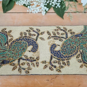 May include: A rectangular, beaded table runner featuring two stylized peacocks. The peacocks are rendered in shades of teal, navy blue, and gold against a cream-colored background. The runner measures approximately 36 inches long.