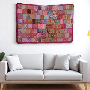 Peut inclure: Une tenture murale patchwork avec une bordure bordeaux, composée de carrés de tissu aux tons roses, oranges et bruns. Au-dessus d'un canapé gris clair avec des coussins blancs. Une lampe de sol jaune est à droite.