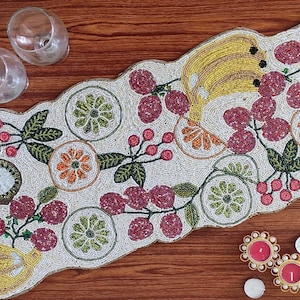 May include: A white beaded table runner featuring a colourful fruit design. The runner depicts bananas, kiwi and berries in shades of red, green and yellow.