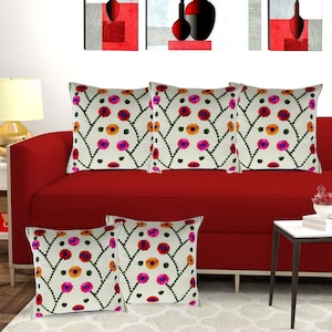 May include: Five white cushion covers with embroidered floral designs in shades of pink, orange, and green. The cushions are arranged on a red sofa.