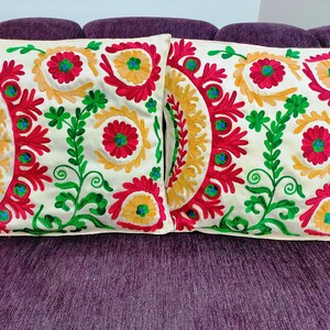 May include: Two embroidered throw cushions with floral designs. The cushions have a cream base, adorned with red, yellow, and green embroidery. The square cushions are placed on a purple sofa, adding a decorative touch.