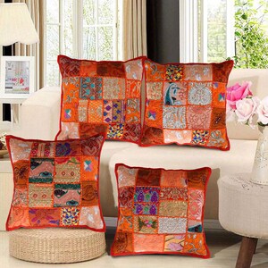 May include: Four square patchwork cushions in orange, red, and gold hues. Each cushion has a red border and a variety of patterned fabric squares. The cushions are arranged on a white sofa and a woven ottoman, creating a warm, inviting aesthetic.
