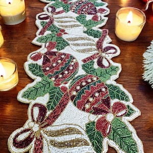 May include: A white beaded table runner with a Christmas theme. The runner features red, green, and gold beads in the shape of baubles, holly leaves, and bows.