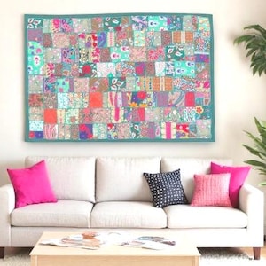 May include: A colorful patchwork quilt with a teal border hangs above a white sofa with pink and black pillows. The quilt features a variety of floral and geometric patterns in shades of pink, teal, orange, and purple. A wooden coffee table sits in front of the sofa.