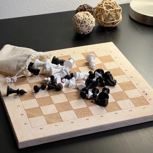 Puede incluir: Un tablero de ajedrez de madera con casillas negras y blancas. Piezas de ajedrez negras y blancas están esparcidas por el tablero, algunas en una bolsa de tela blanca. El tablero tiene números y letras impresos en los lados.