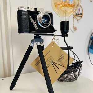 Könnte beinhalten: Eine Vintage-Kamera auf einem Stativ mit einer Glühbirne an der Seite. Die Kamera ist schwarz und silber mit einem Objektiv vorne. Die Glühbirne ist aus klarem Glas mit einem Filament im Inneren. Das Stativ ist aus schwarzem Metall mit drei Beinen. Die Glühbirne hat ein Etikett mit der Aufschrift "home".