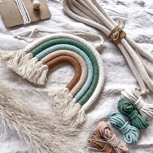 Könnte beinhalten: Ein Regenbogen-Makramee-Wandbehang mit einem weißen Quasten. Der Regenbogen besteht aus fünf verschiedenen Farben von Seil: Braun, Rosa, Hellblau, Grün und Dunkelgrün. Das Seil ist oben am Regenbogen zu einem Knoten gebunden. Es gibt auch fünf Spulen mit Seil in verschiedenen Farben: Braun, Rosa, Hellblau, Grün und Dunkelgrün. Die Spulen mit Seil befinden sich auf einem weißen Leinenhintergrund.