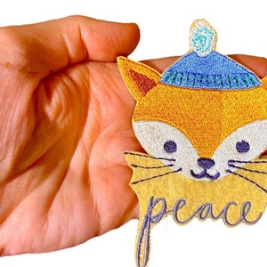May include: Embroidered iron-on patch featuring a fox wearing a blue and white knit hat with a white pom pom. The fox has a yellow body, white face, and purple eyes. The patch has a yellow background with the word "peace" embroidered in purple.
