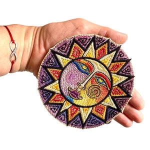 Könnte beinhalten: Ein runder gestickter Patch mit einem farbenfrohen Sonnen-Gesichtsdesign. Die Sonne hat ein lila, gelbes und blaues Gesicht mit einem roten Mund und gelben Augen. Die Sonne ist von einem lila, gelben und roten Mandala-Design umgeben.