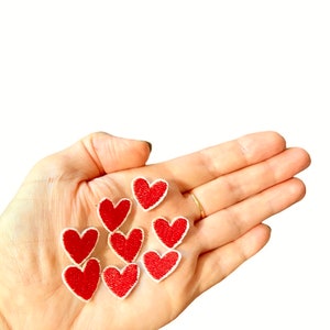 May include: A hand holding eight red felt heart patches with white outlines. The patches are small and perfect for adding a touch of love to crafts and projects.