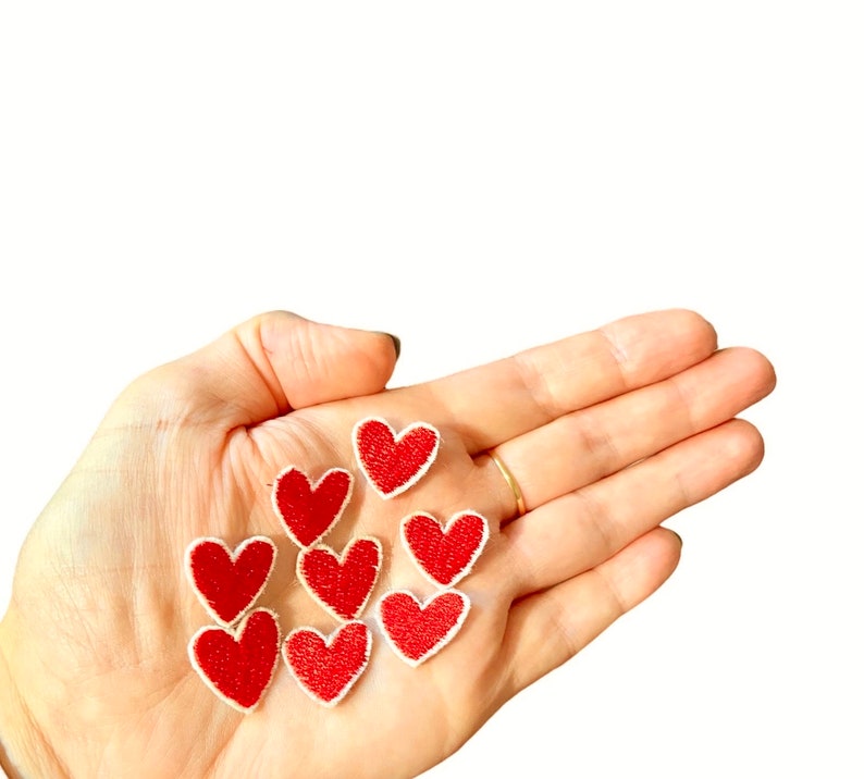May include: A hand holding eight red felt heart patches with white outlines. The patches are small and perfect for adding a touch of love to crafts and projects.