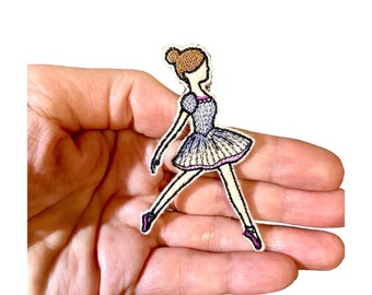 May include: Embroidered patch of a ballerina in a graceful pose. The patch features a light purple tutu, a bun, and pink ballet slippers. The design is outlined in black thread, creating a detailed and elegant look.