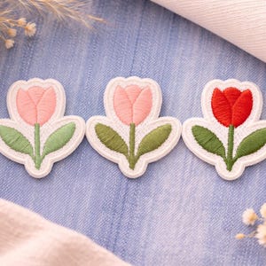 Könnte beinhalten: Drei gestickte Tulpenaufnäher in Rosa und Rot mit grünen Blättern und Stielen. Die Aufnäher haben einen weißen Rand und sind auf einem blauen Jeans-Hintergrund angeordnet. Dekorative florale Elemente im Hintergrund.