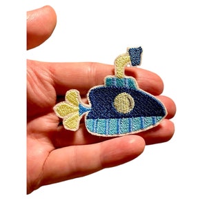Puede incluir: Parche bordado de un submarino azul con periscopio y hélice amarillos. El submarino tiene una franja azul claro y un ojo de buey amarillo. El parche se sostiene en una mano.
