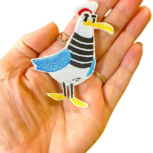 May include: A white and blue embroidered iron-on patch of a cartoon seagull wearing a red and white sailor hat with a black stripe. The seagull has black eyes, a yellow beak, and yellow feet.