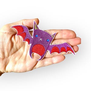 May include: Purple and red embroidered iron-on patch of a cartoon bat with big eyes.