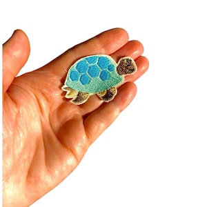 May include: Embroidered turtle patch with a blue and green shell, brown head and flippers. The patch is held in a hand against a white background. The turtle patch is approximately 5 cm long.