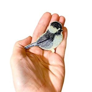 Puede incluir: Un parche bordado de un pájaro negro y blanco con un contorno amarillo. El pájaro está posado en una rama y tiene una cabeza pequeña y redonda.