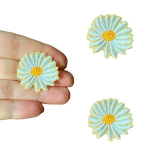 Op de afbeelding: Drie lichtblauwe geborduurde madeliefjes met gele centra. De madeliefjes zijn op een witte achtergrond.