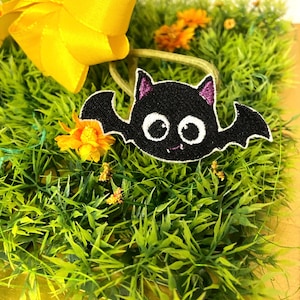 May include: A black bat with white eyes and purple ears is sitting on a bed of green grass. The bat is surrounded by yellow flowers.