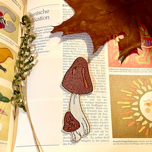 Könnte beinhalten: Ein gestickter brauner und weißer Pilz-Patch mit einem weißen Stiel und einer braunen Kappe. Der Patch befindet sich auf einer Buchseite mit deutschem Text über Indianertechnik.