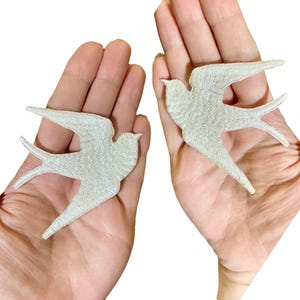 Puede incluir: Dos parches bordados de golondrina blanca. Los parches se muestran en manos, con los pájaros mirando en direcciones opuestas.