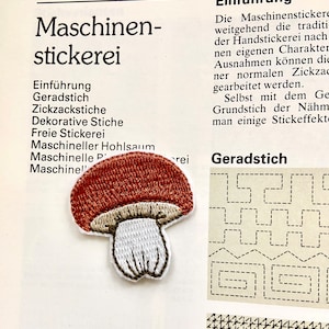 Op de afbeelding: Een close-up van een bruin en wit gestikt patch in de vorm van een paddenstoel. Het patch bevindt zich op een witte achtergrond met tekst over machineborduren.