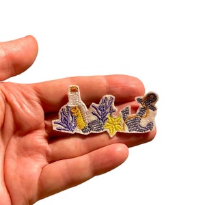 Può includere: Toppa ricamata a tema nautico, raffigurante una bottiglia, un'ancora e del fogliame. La toppa utilizza fili bianchi, viola, gialli e dorati. Il design presenta cuciture dettagliate e un aspetto strutturato. Destinata all'applicazione su abbigliamento o accessori.