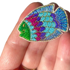 Peut inclure: Un patch brodé de poisson coloré avec du fil vert, bleu, violet et or. Le patch est sur un fond blanc et mesure environ 5 cm de long.