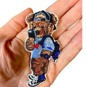 Op de afbeelding: Gestikt patch van een bruine beer die een blauw shirt, een blauwe spijkerbroek, een blauwe rugzak en een blauwe baseballpet draagt. De beer houdt een zwarte telefoon in zijn rechterpoot.