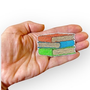 Peut inclure: Un ensemble de trois patchs brodés thermocollants représentant une pile de livres. Le livre du haut est orange, le livre du milieu est bleu et le livre du bas est vert.