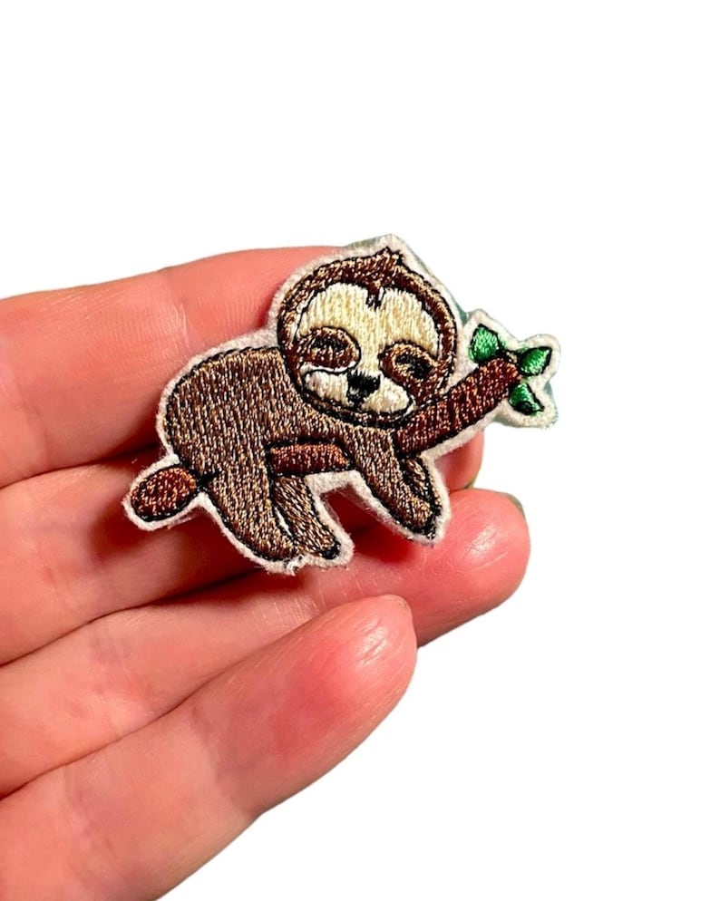 K&ouml;nnte beinhalten: Gestickter B&uuml;gel-Patch mit einem braunen Faultier mit einem schl&auml;frigen Ausdruck, das an einem braunen Ast mit gr&uuml;nen Bl&auml;ttern h&auml;ngt.