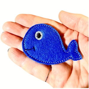 Pode incluir: Uma baleia de feltro azul com costuras brancas e um olho preto. A baleia tem aproximadamente 10 cm de comprimento e é ideal para um brinquedo infantil ou um acento decorativo.