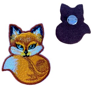 Könnte beinhalten: Gestickter Flicken mit einem Fuchsmotiv in Orange, Weiß und Blau. Der Flicken zeigt ein detailliertes Design eines Fuchses mit eingerolltem Schwanz. Die Rückseite des Flicken ist dunkelbraun und hat eine Anstecknadel. Ein dekoratives Accessoire.