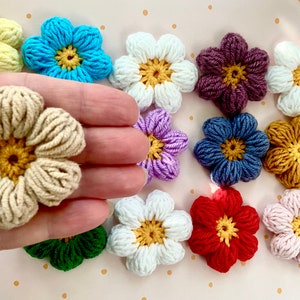 May include: A collection of crocheted flower embellishments in various colours, including yellow, blue, white, purple, red, green, brown, and pink. The flowers have a five-petal design with a yellow centre.