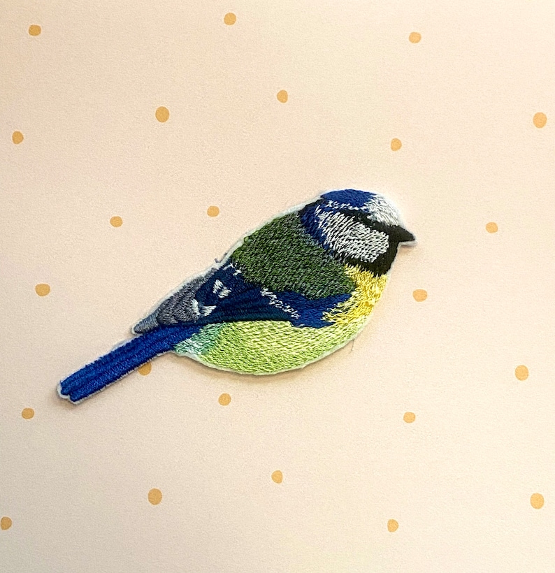 May include: Embroidered bluebird patch with a blue, green, and yellow design. The bird is facing right and has a small, pointed beak.